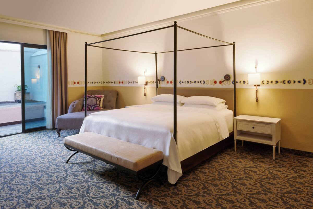 Ein Hotelzimmer mit Himmelbett, einer Sitzbank, einem Nachttisch und Blick ins Freie.
