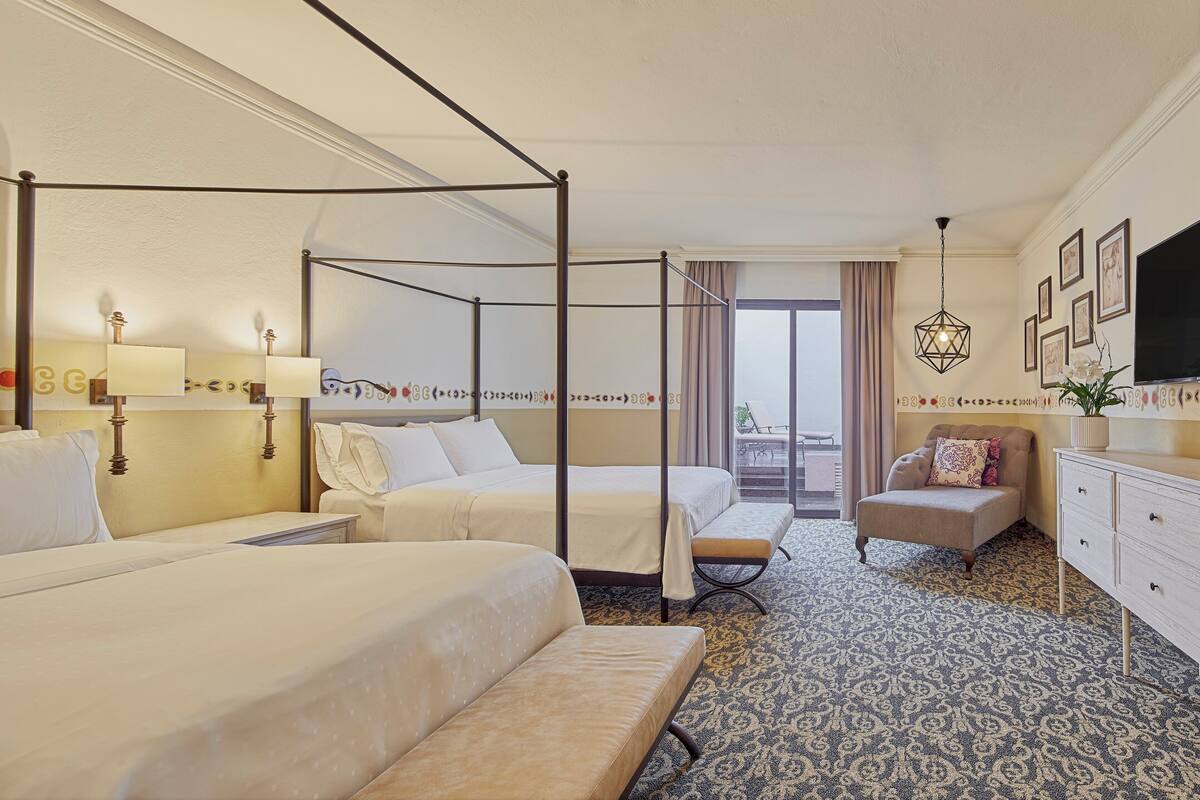 Ein Hotelzimmer mit zwei Betten, einem Sofa, einem Fernseher und einem Balkon mit Vorhängen.