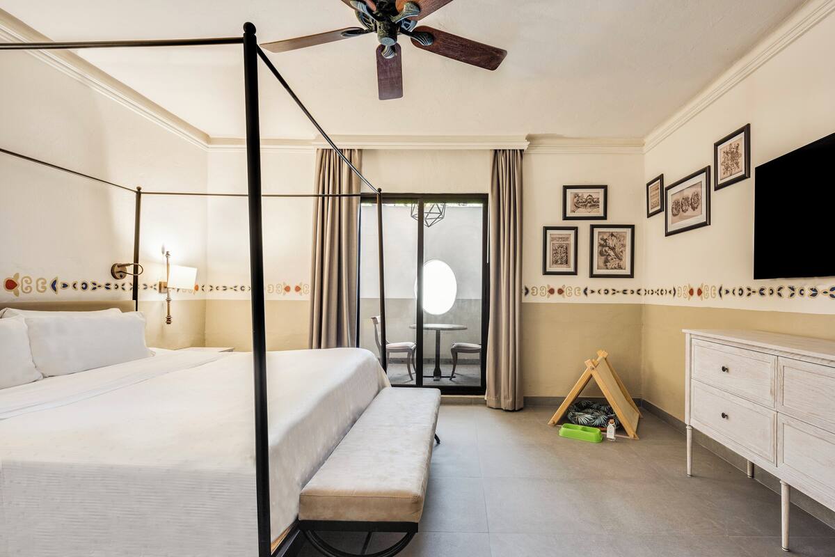 Hotelkamer met een hemelbed, een bankje, een plafondventilator, een televisie, een zithoek met een tafel en stoelen, en een kleine kinderspeelruimte met een tent en speelgoed.