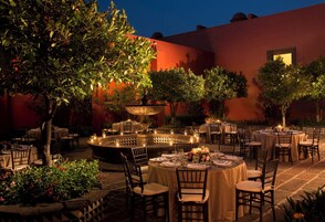 An outdoor dining area with round tables and chairs, surrounded by trees and a decorative fountain.