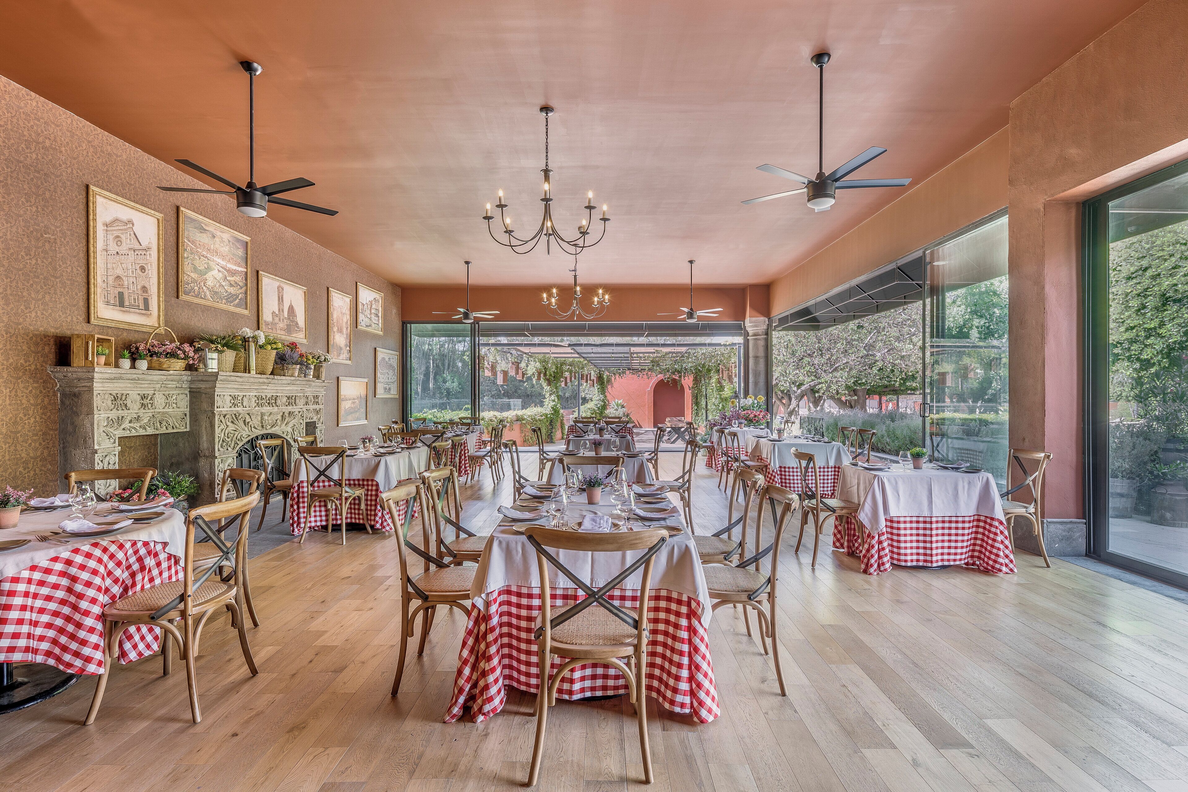 A restaurant with high ceilings, chandeliers, and large windows offering a view of the outdoors. Tables are set with red and white checkered tablecloths, and the room has a warm, rustic wooden floor.