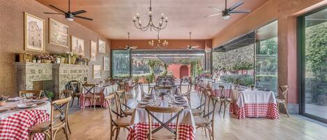 A restaurant with high ceilings, chandeliers, and large windows offering a view of the outdoors. Tables are set with red and white checkered tablecloths, and the room has a warm, rustic wooden floor.