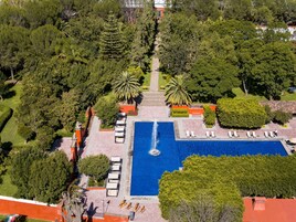 Vista aérea de un complejo turístico con piscina central, rodeado de exuberante vegetación y palmeras.