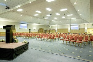Un salón de conferencias amplio con filas de sillas, un escenario con un atril y dos pantallas.
