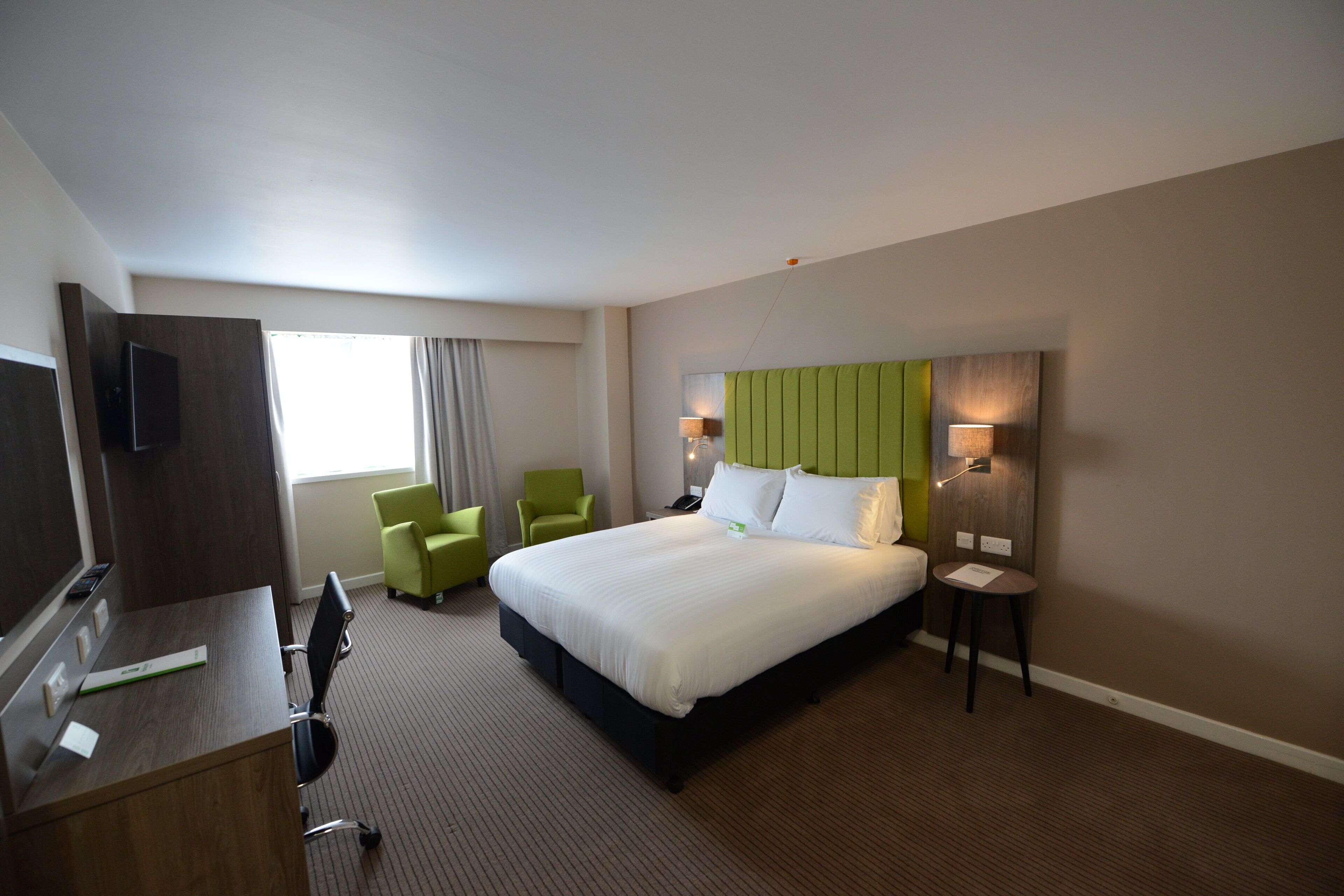Quarto de hotel com cama, duas cadeiras, escrivaninha e televisão.
