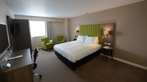Habitación de hotel con una cama grande, un escritorio con silla, un sillón verde y una mesita de noche con lámpara.