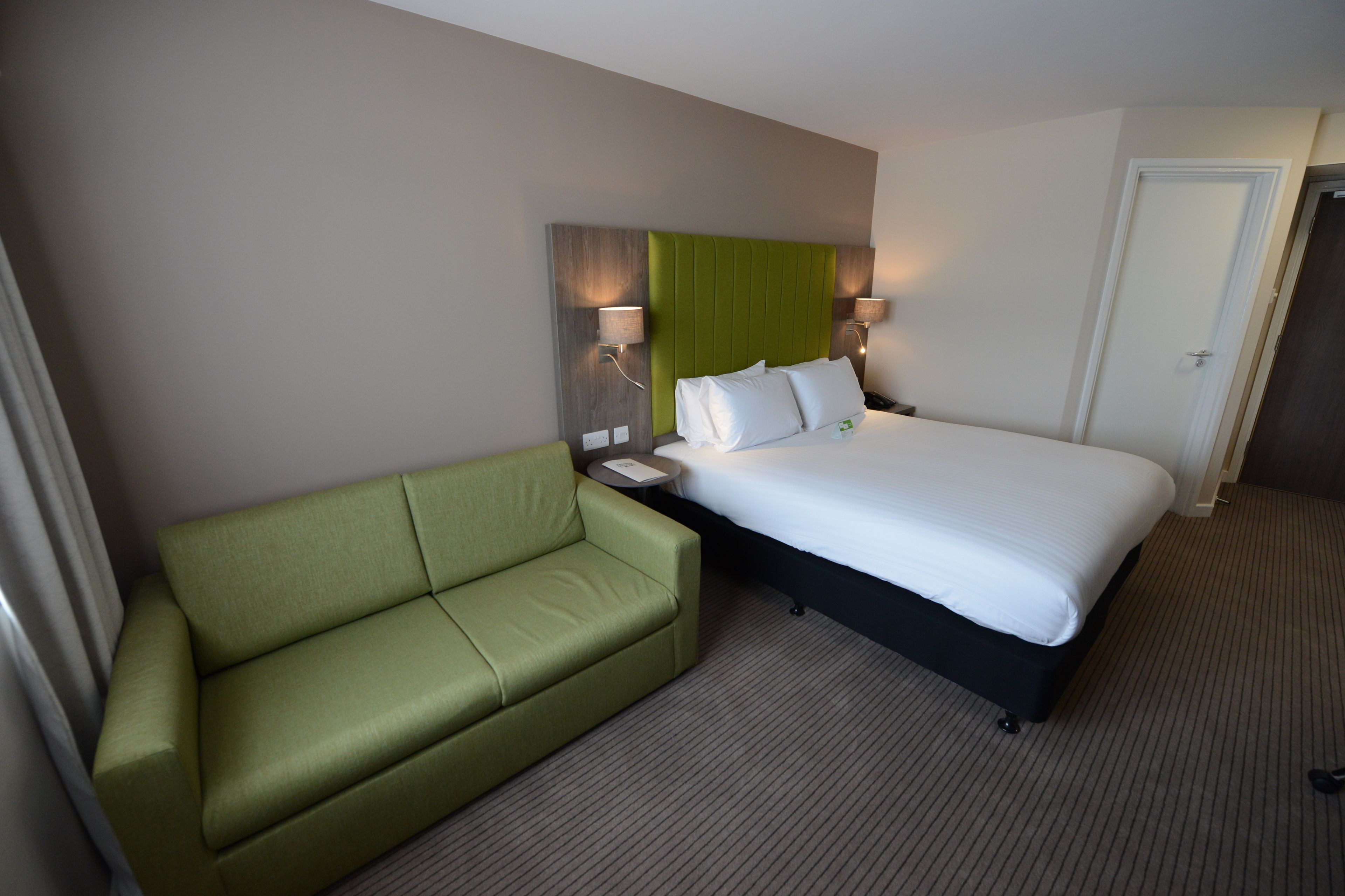 A hotel room with a bed, a green sofa, two bedside lamps, and a door.