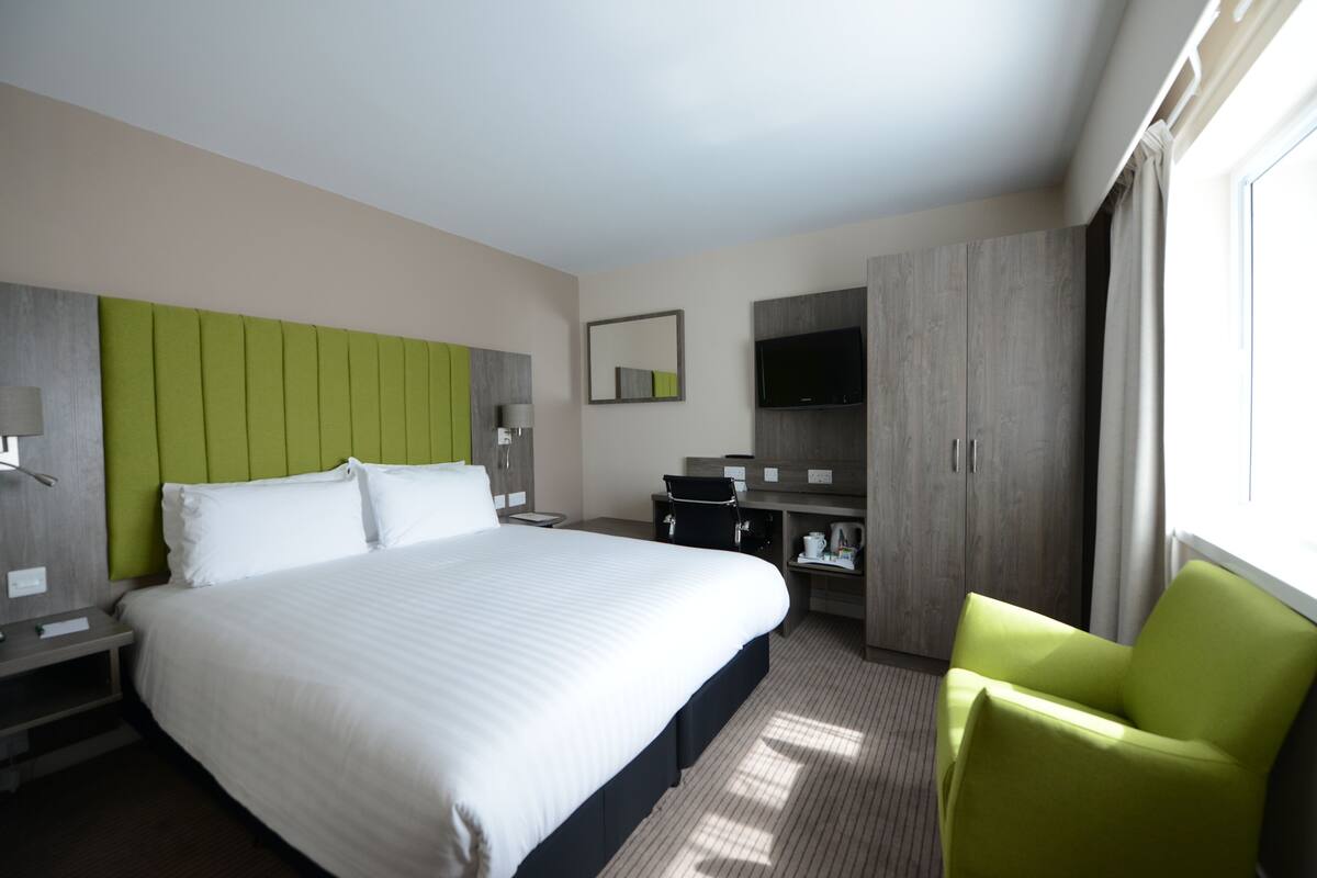 A hotel room with a large bed, a green upholstered chair, a flat-screen TV, a wardrobe, and a desk with a lamp.