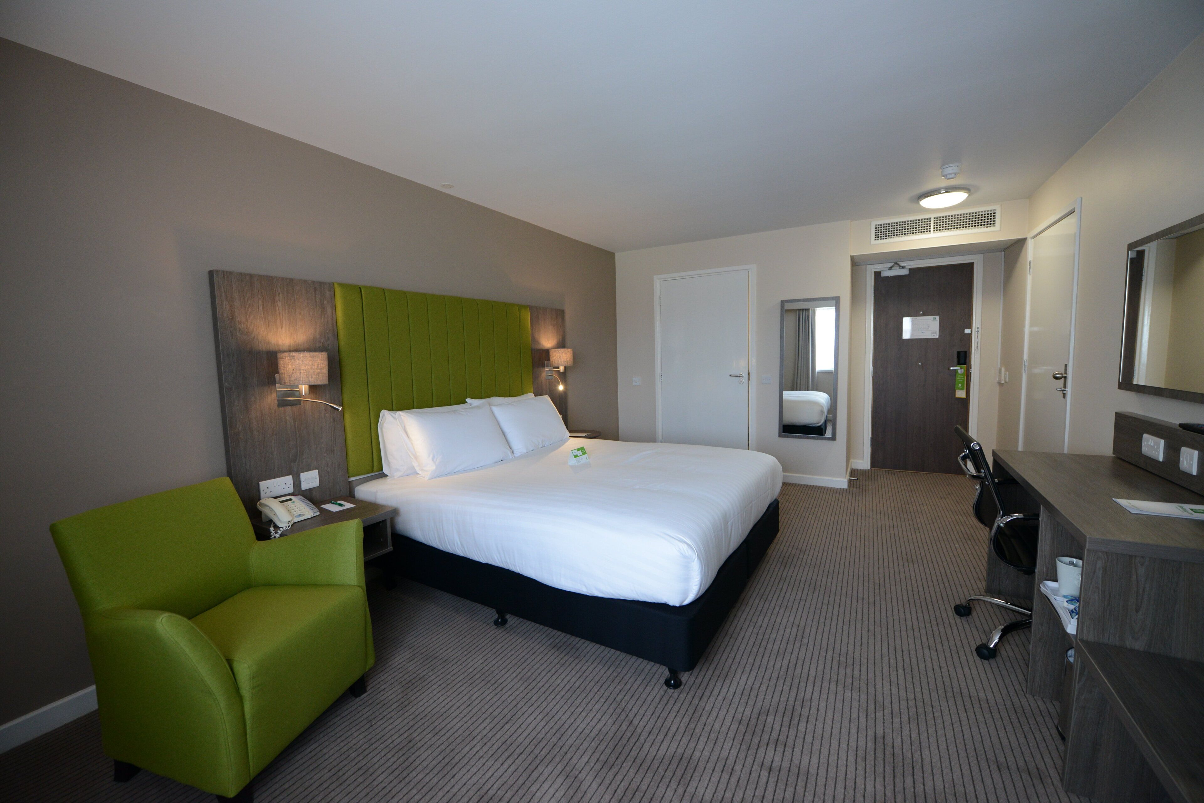 A hotel room with a bed, a green armchair, a desk with a telephone, and a mirror.