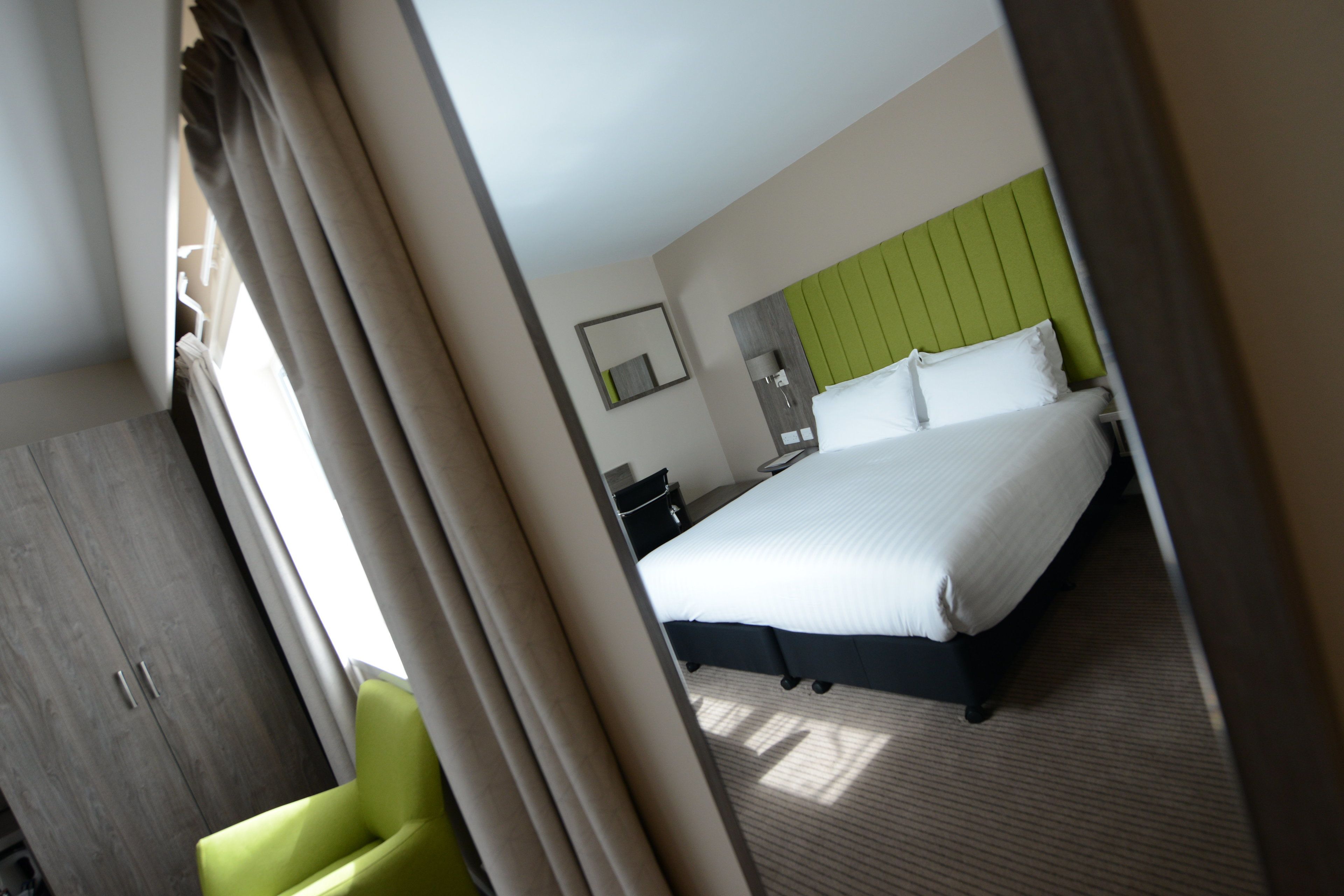 A hotel room with a bed, a green headboard, a nightstand, a mirror, and a wardrobe.