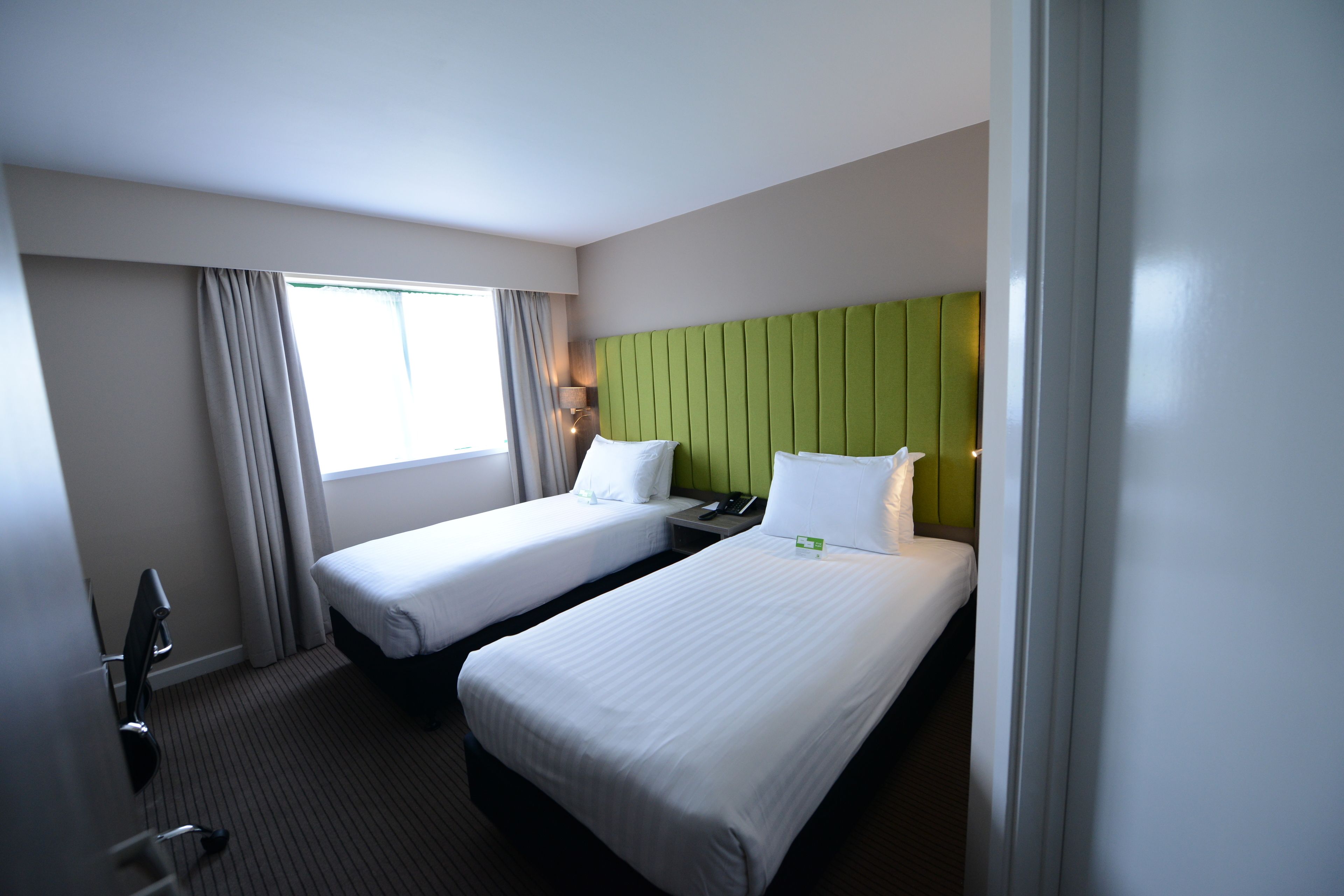 Una habitación de hotel con dos camas individuales, un cabecero verde, una mesita de noche con lámpara y una ventana con cortinas.