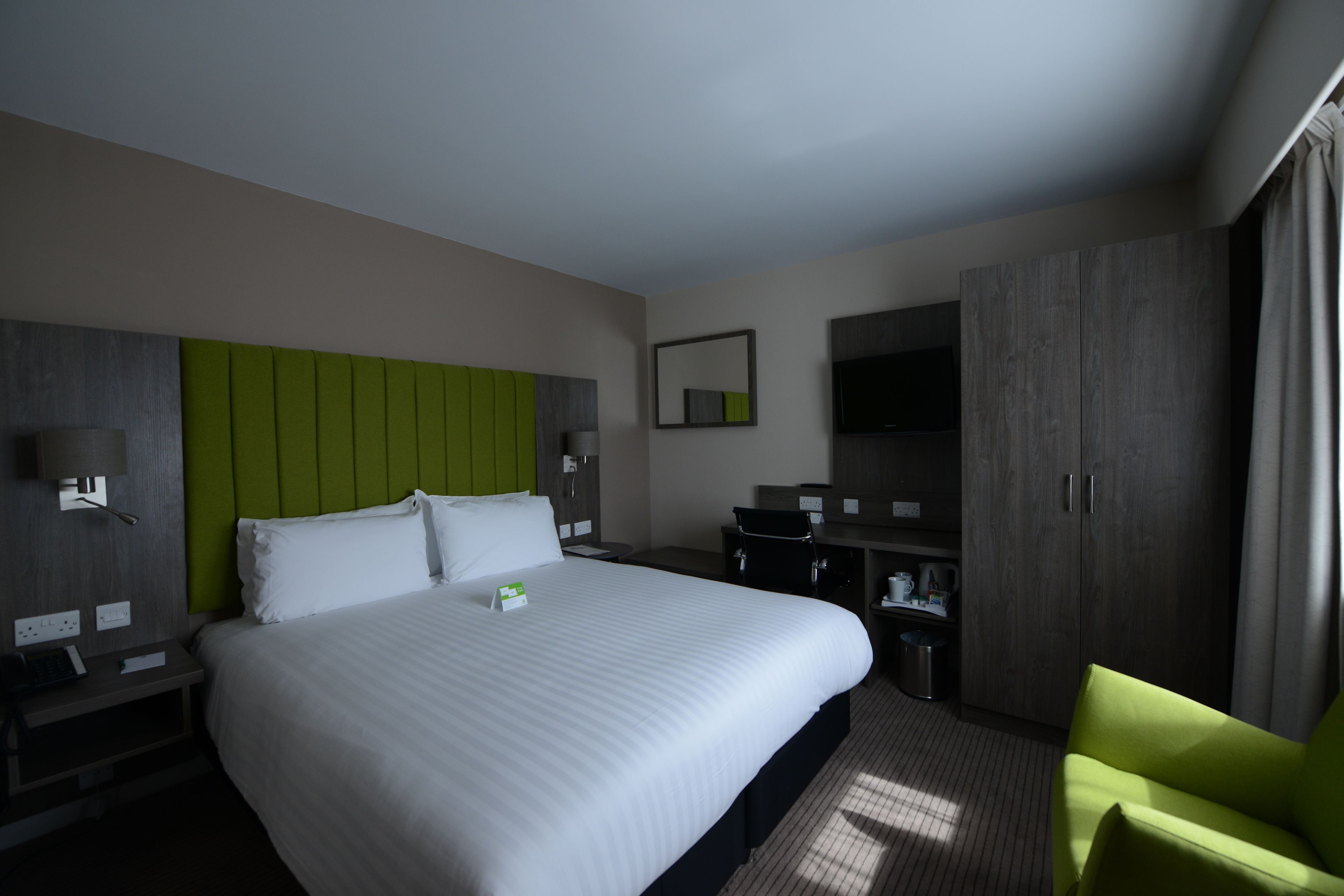 A hotel room with a large bed, a green accent wall, a desk with a chair, a TV, and a wardrobe.