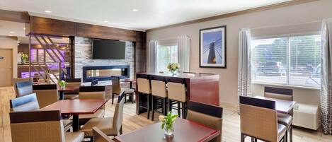 A modern dining area with wooden tables and cushioned chairs, a flat-screen TV mounted on the wall, and a staircase leading to an upper level.