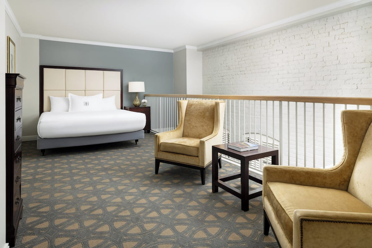 Quarto de hotel com cama, duas poltronas, uma mesinha e uma parede de tijolos brancos.