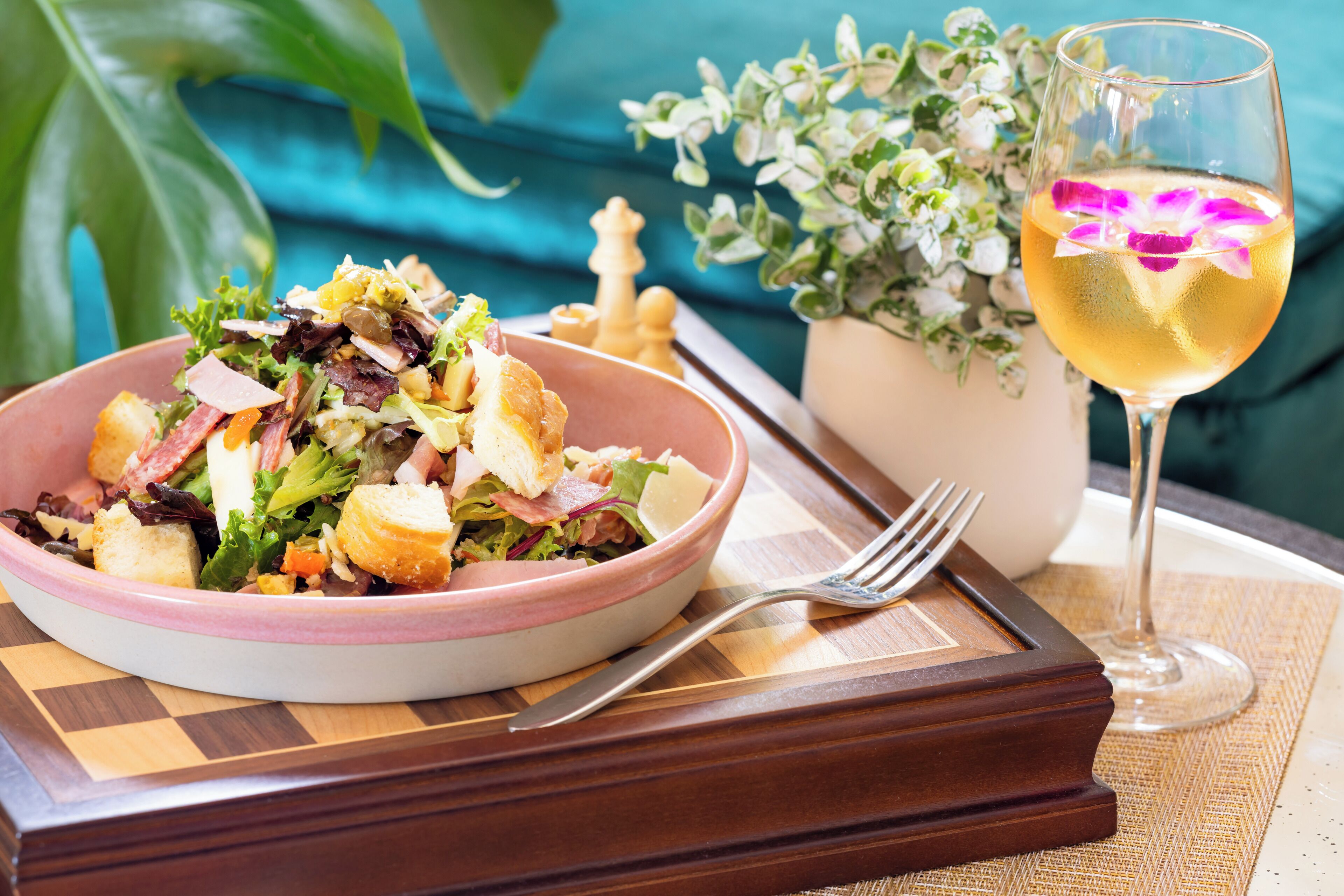 Un saladier rempli de salade, avec des croûtons et du fromage, un verre de vin blanc orné d’une fleur violette, et un jeu d’échecs avec les pièces.