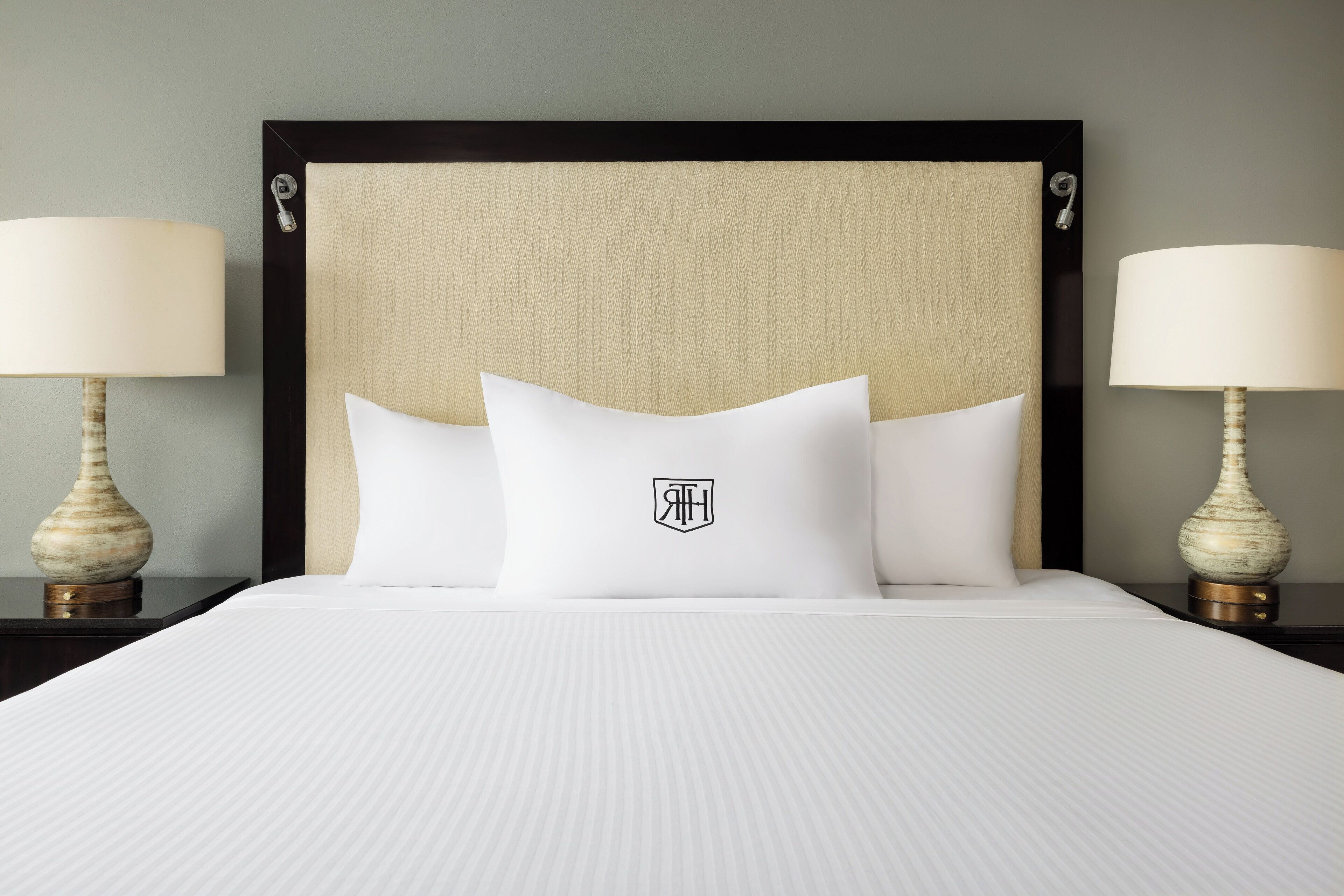Una cama bien hecha con cabecera, dos lámparas de noche y un logotipo en la almohada.