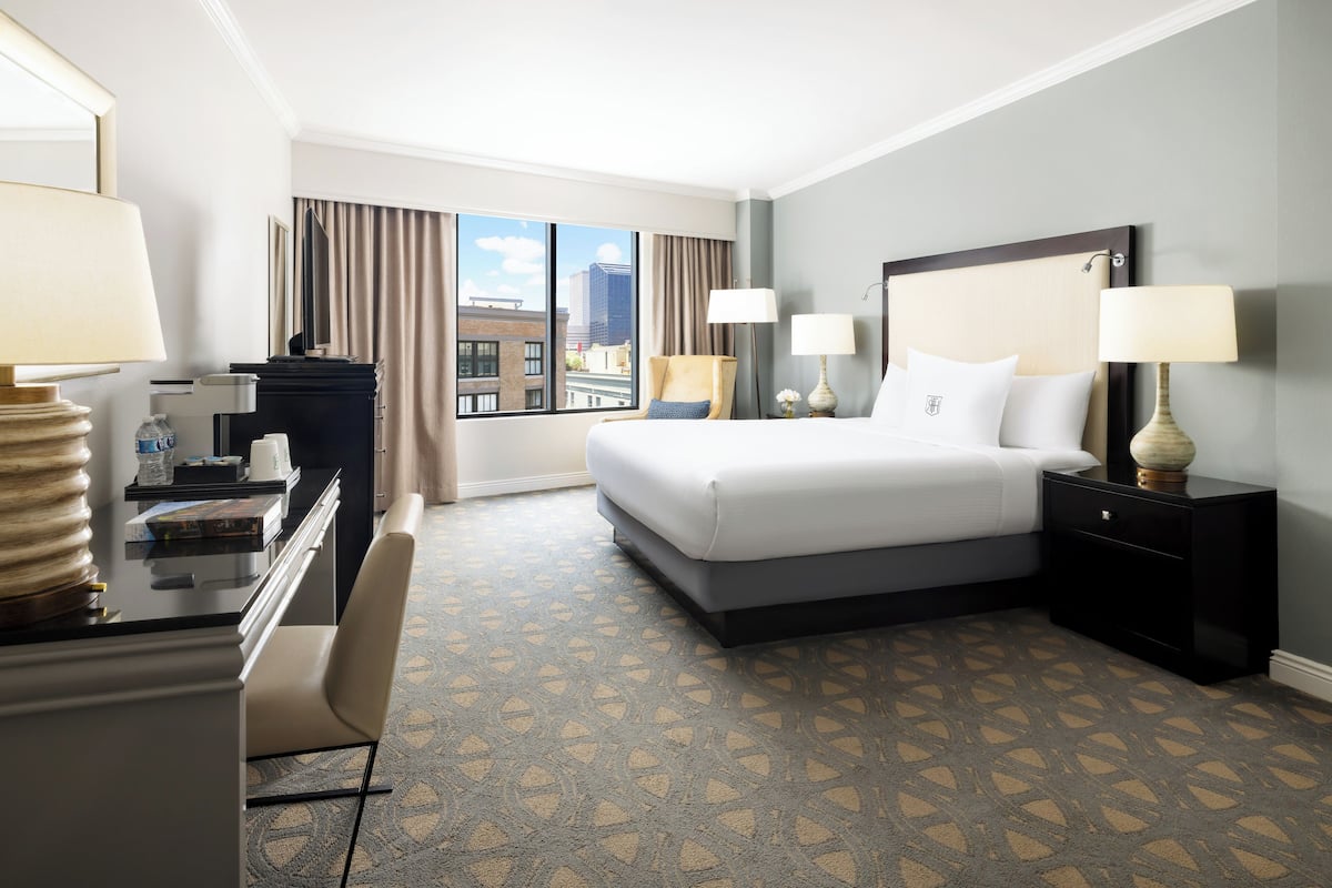 Quarto de hotel com uma cama grande, duas mesas de cabeceira com abajures, uma escrivaninha com cadeira e vista para a cidade pela janela.