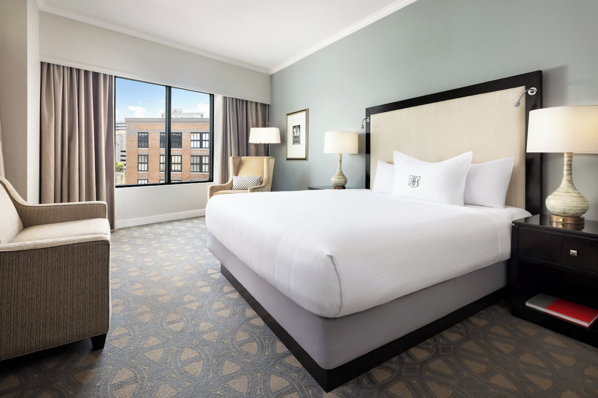 Quarto de hotel com uma cama grande, uma poltrona, uma mesa de cabeceira e vista para um edifício pela janela.