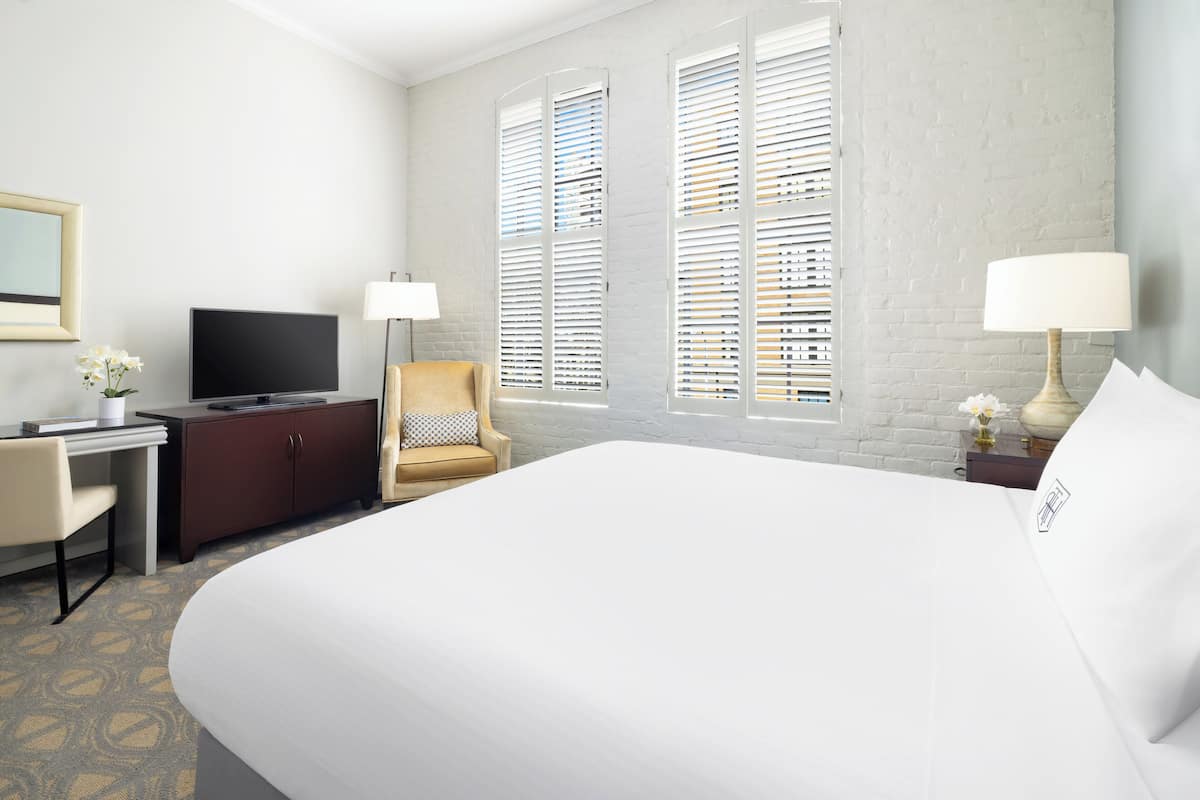 Quarto de hotel com uma cama grande, uma escrivaninha com televisão, uma cadeira e duas janelas com persianas.