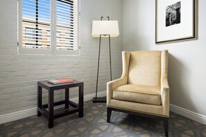 Ein Zimmer mit einem beigefarbenen Sessel, einem kleinen Holztisch mit einem Buch, einer Stehlampe und einem Fenster mit Jalousien.