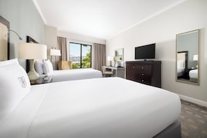 A hotel room with two beds, a desk, a television, and a large window with a view.