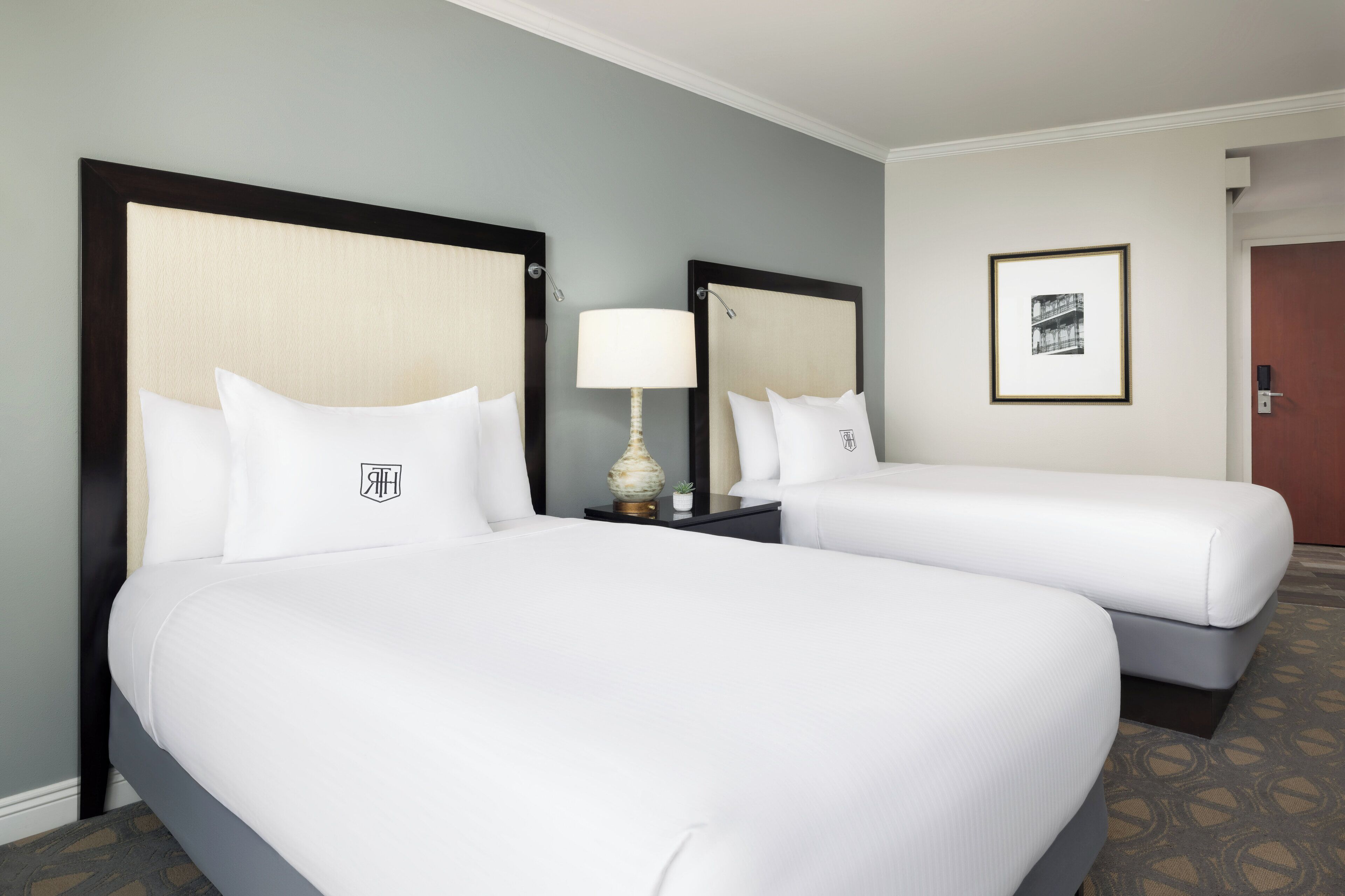 A hotel room with two beds, a nightstand with a lamp, a framed picture on the wall, and a door to the right.