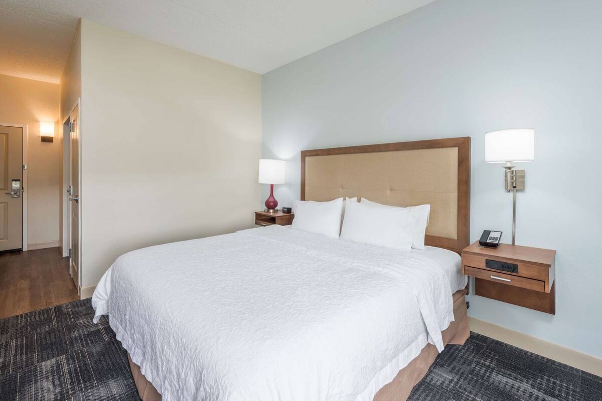 A hotel room with a large bed, a nightstand with a lamp, a bedside table with a phone, and a door leading to another room.