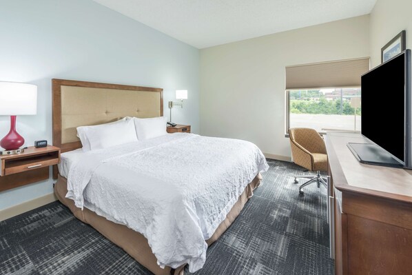 A hotel room with a large bed, a desk with a computer, a chair, a TV, and a window with a view of the outside.