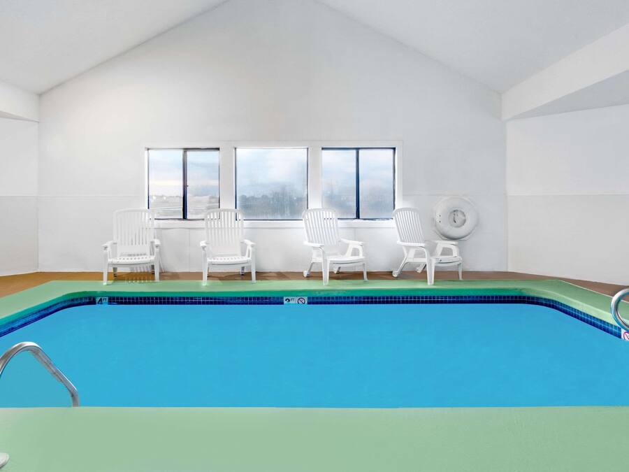 Piscina com água azul translúcida, rodeada por cadeiras brancas, e grandes janelas com vista para o céu.