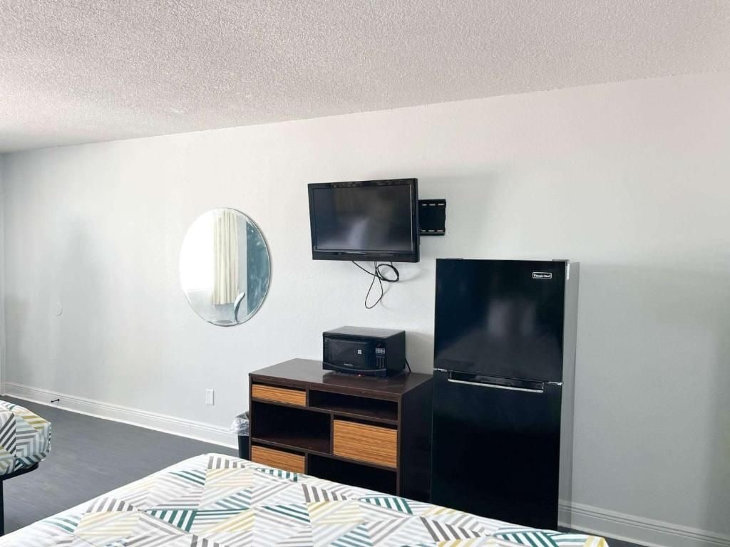Ein Hotelzimmer mit einem Bett, einem flachbildfernseher an der Wand, einem Kühlschrank, einem kleinen Nachttisch und einem Spiegel.