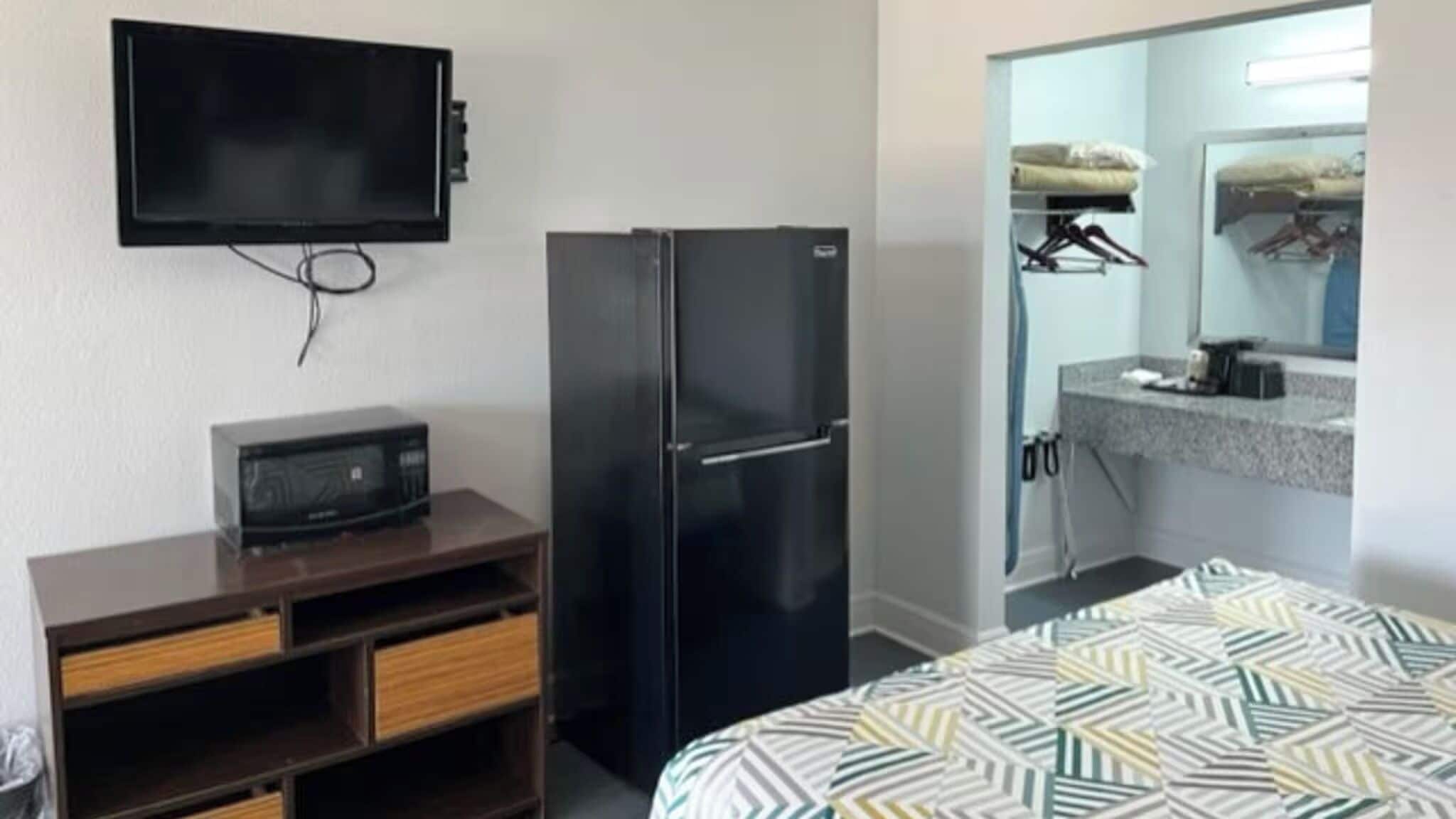 A hotel room with a bed, a flat-screen TV mounted on the wall, a refrigerator, a microwave, a wooden dresser with drawers, and a bathroom with a sink and mirror.