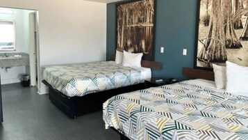 Quarto de hotel com duas camas, uma mesinha de cabeceira, banheiro com pia e um quadro abstrato grande na parede.