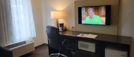 A hotel room with a desk, chair, air conditioning unit, lamp, television, and a mini-fridge.