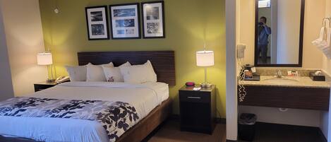 A hotel room with a bed, bedside lamps, a nightstand, a mirror, a vanity with a sink, and framed pictures on the wall.