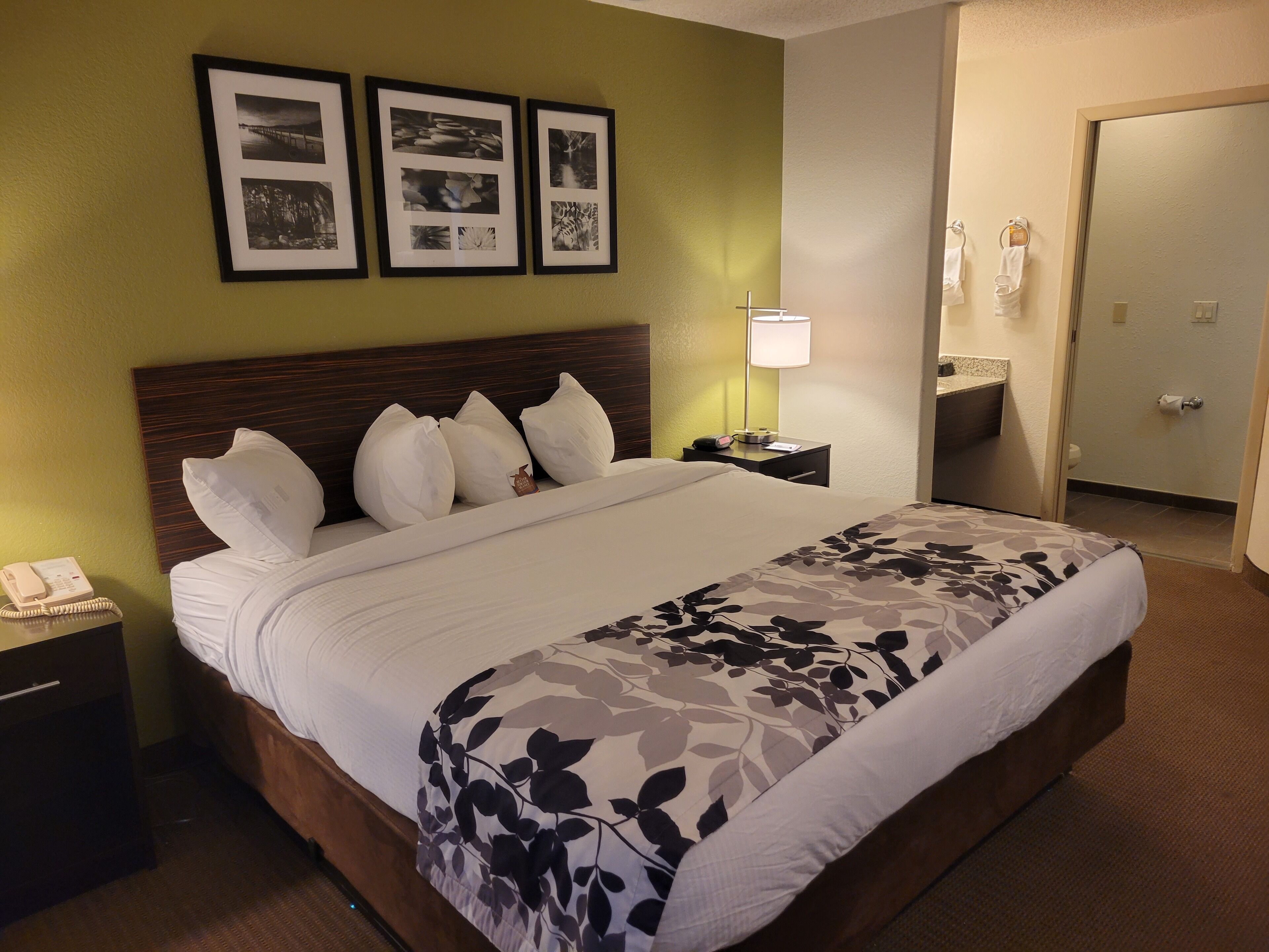 A hotel room with a large bed, a nightstand with a phone, a lamp, and a framed photo collage on the wall.