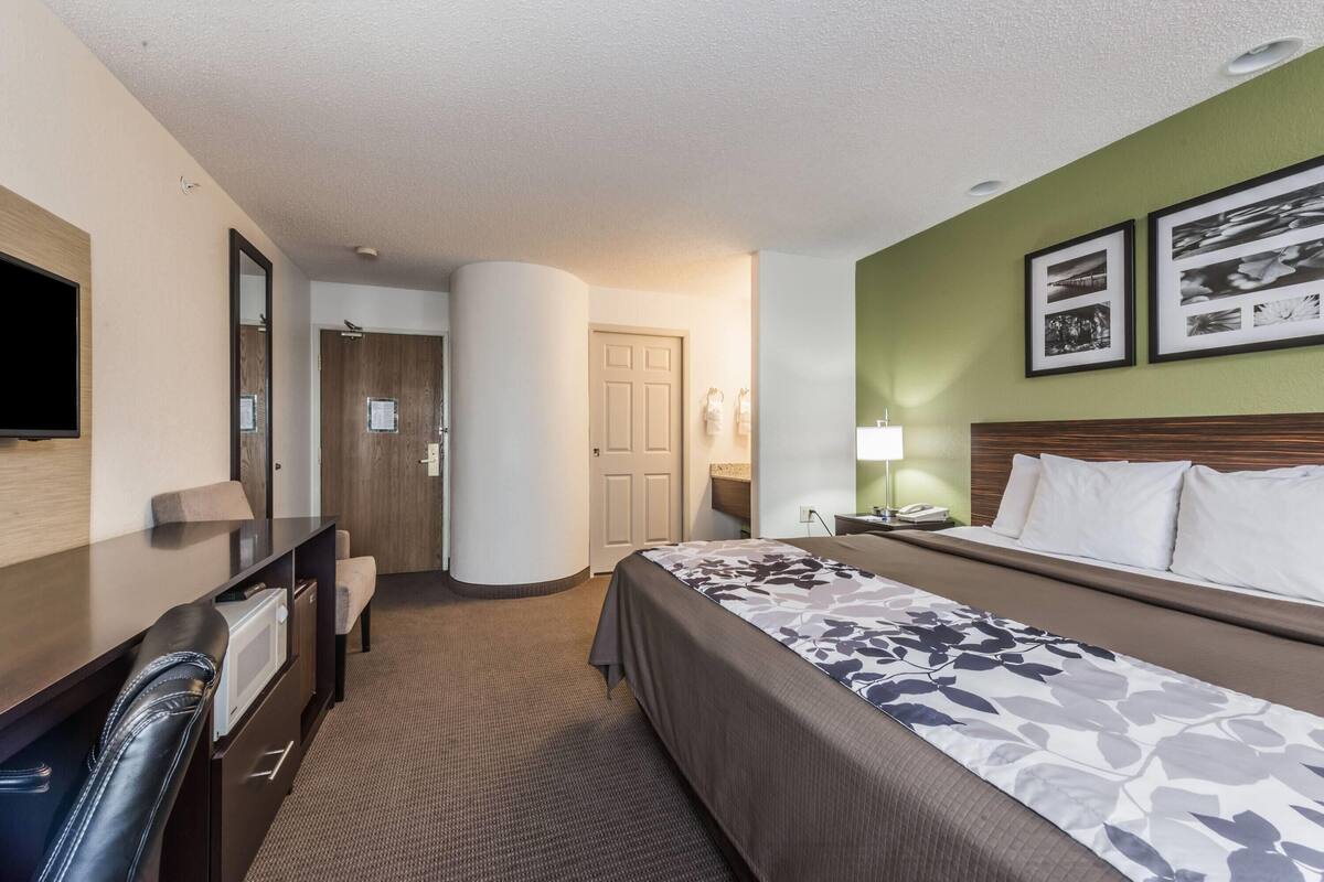 Ein Hotelzimmer mit einem Bett, einem Schreibtisch, einem Stuhl, einem Fernseher, einem Spiegel und gerahmten Bildern an der Wand.