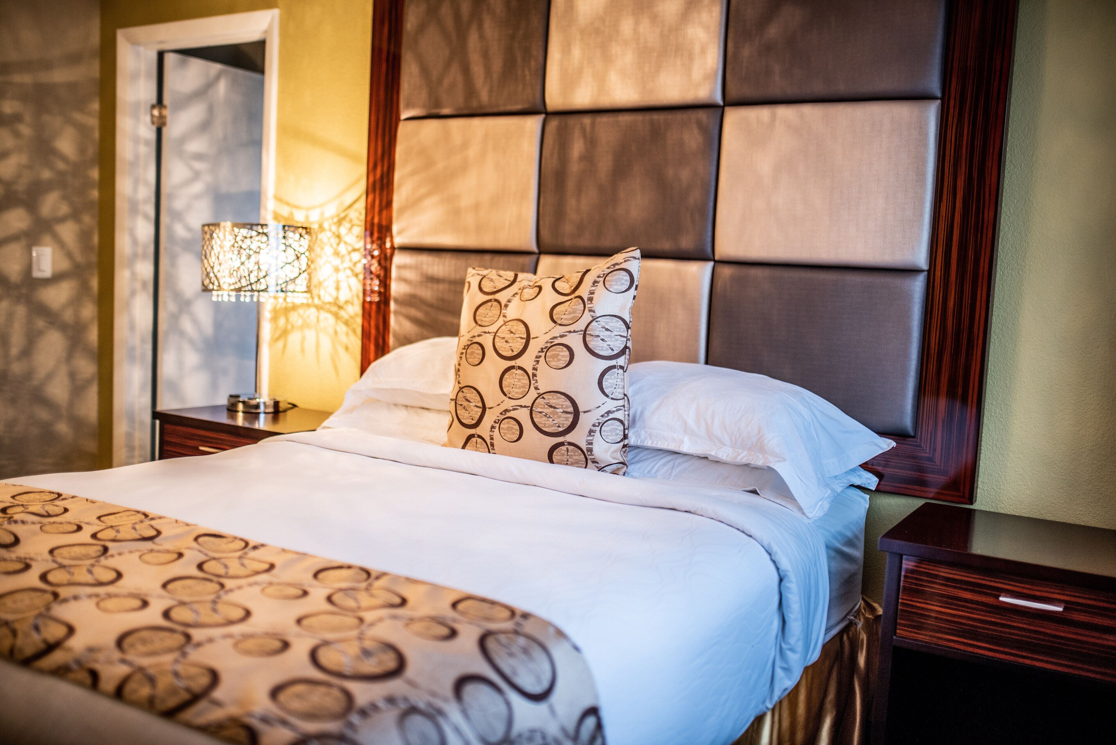 A neatly made bed with patterned bedding, a headboard with a padded design, and a bedside lamp.