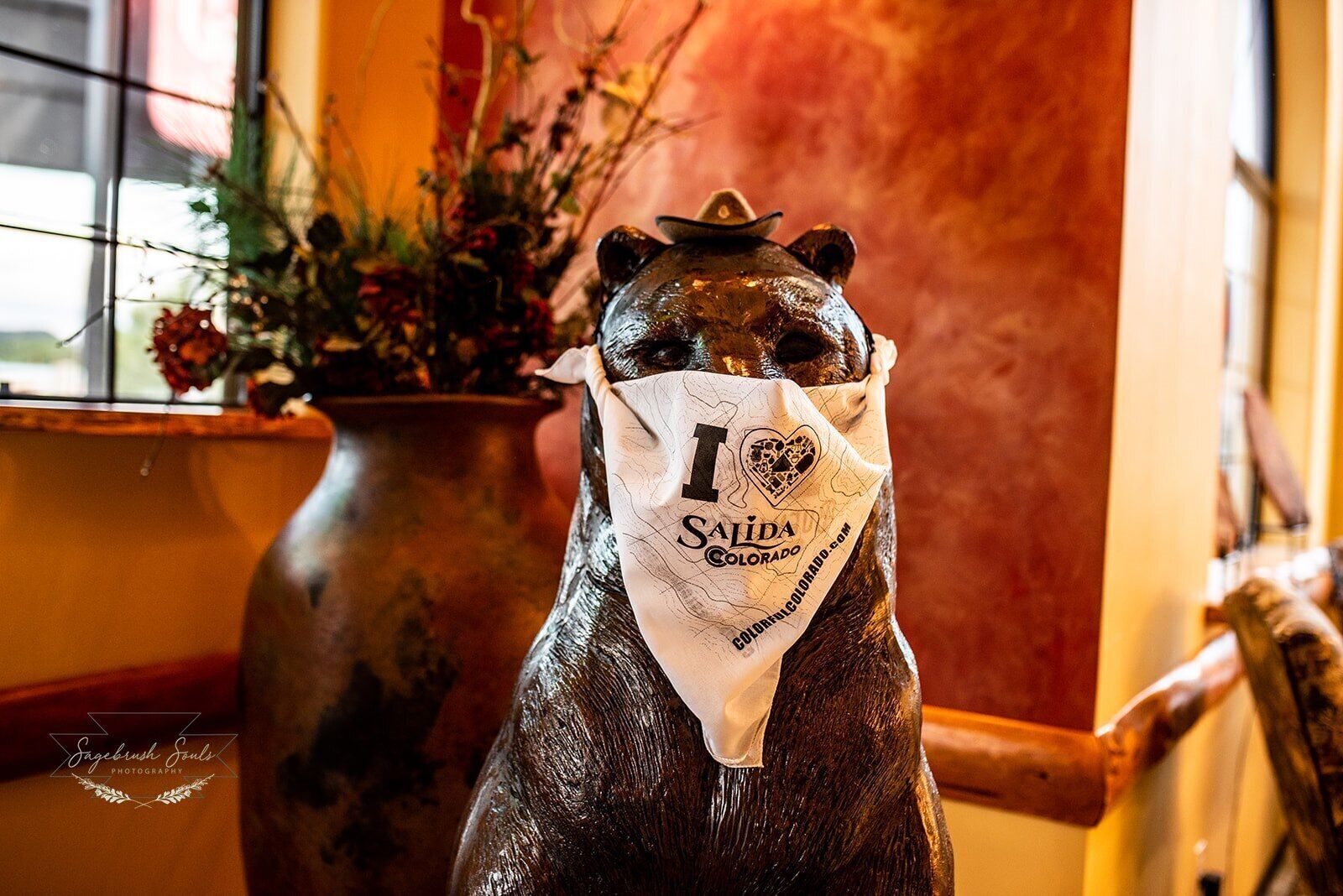 En bjørnestatue med bandana og teksten "1 Salida Colorado", placeret i et rum med en blomstervase og en træstol.