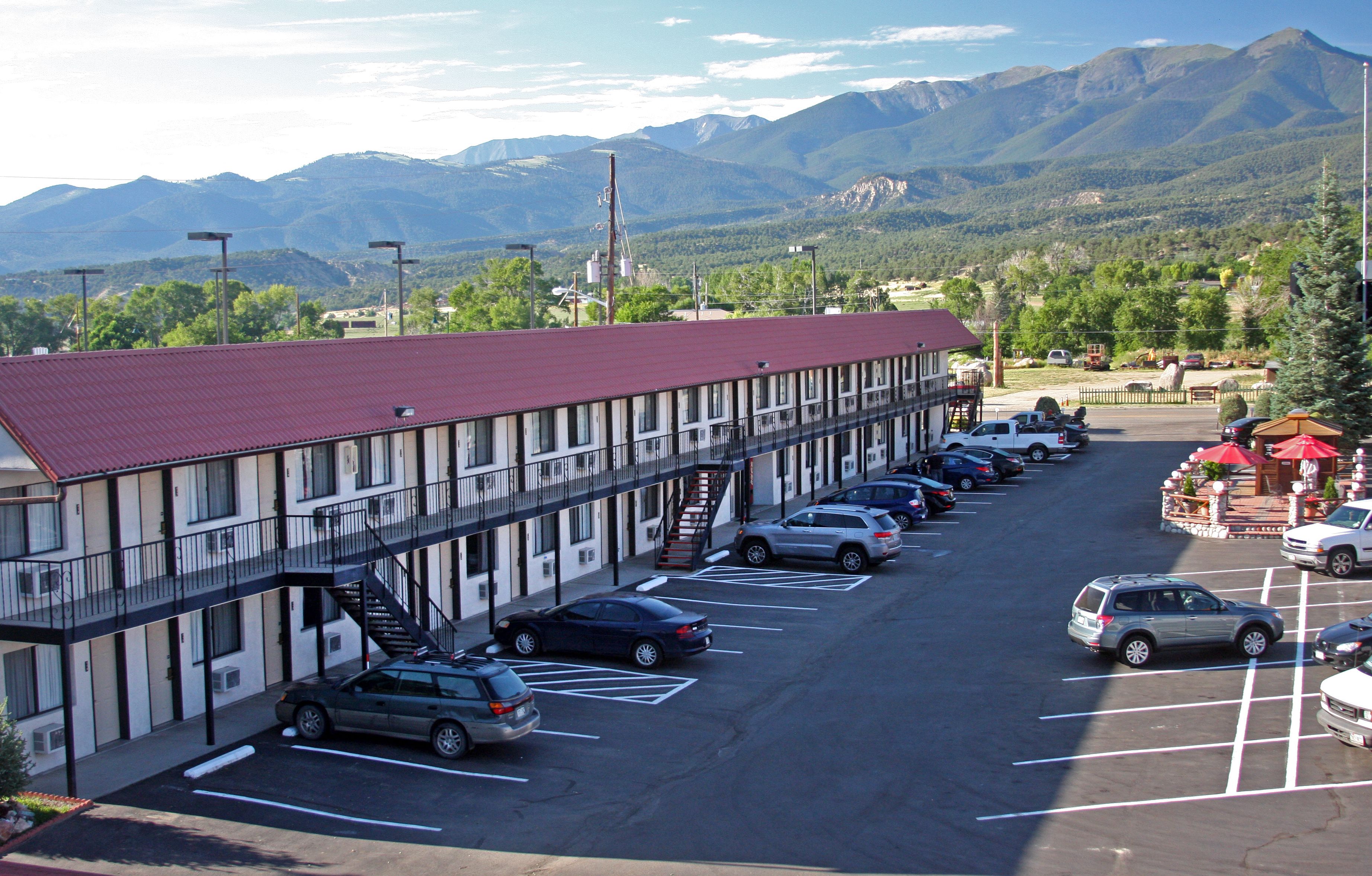 A motel with a red roof, multiple parking spaces, and a view of mountains in the background.