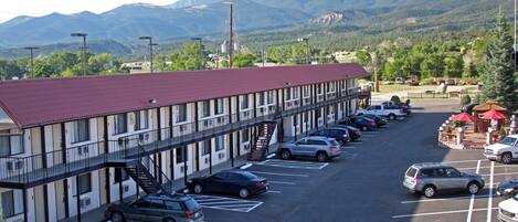 A motel with a red roof, multiple parking spaces, and a view of mountains in the background.