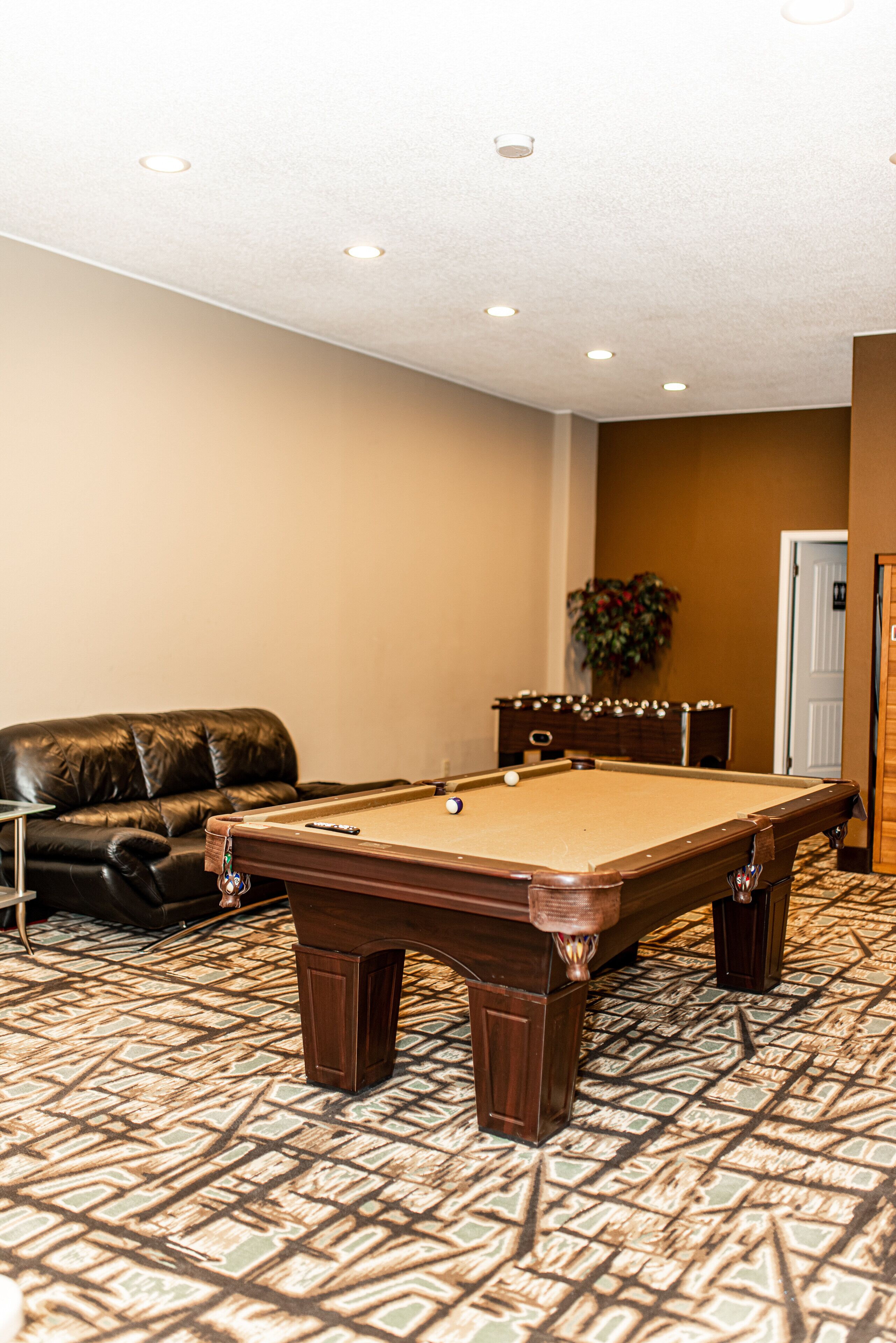 A pool table in a room with a patterned carpet, a leather couch, and a door in the background.