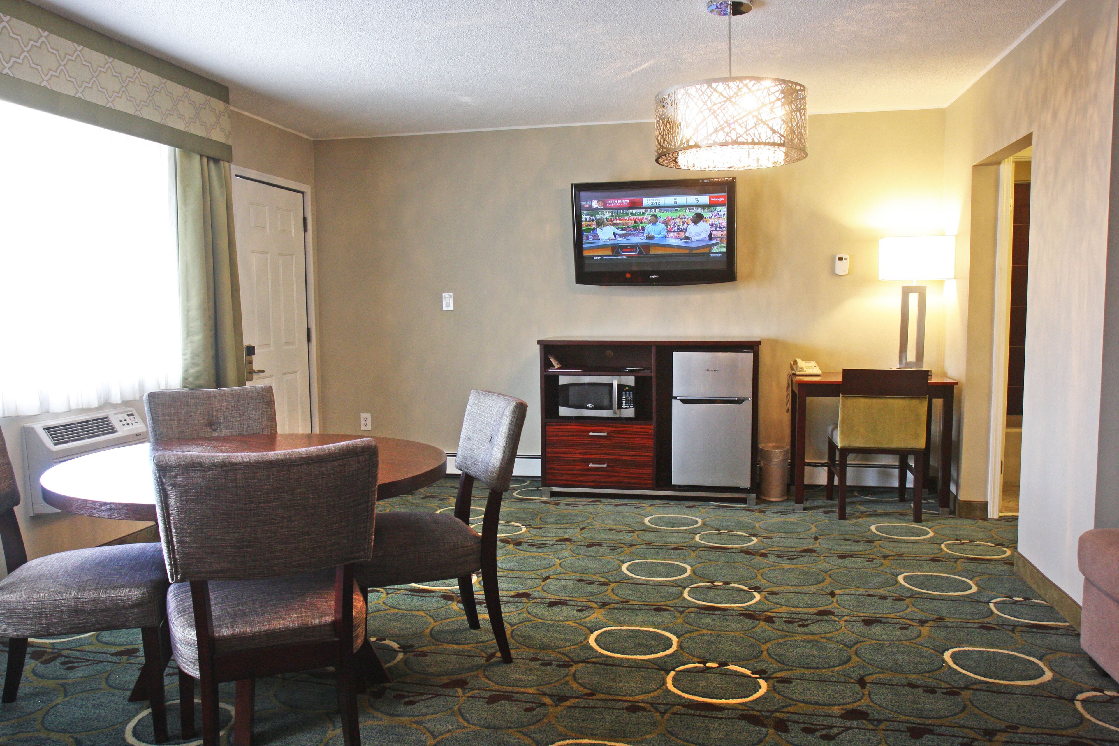 A hotel room with a dining area, a television mounted on the wall, a microwave, a refrigerator, and a lamp.