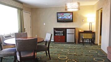 A hotel room with a dining area, a television mounted on the wall, a microwave, a refrigerator, and a lamp.