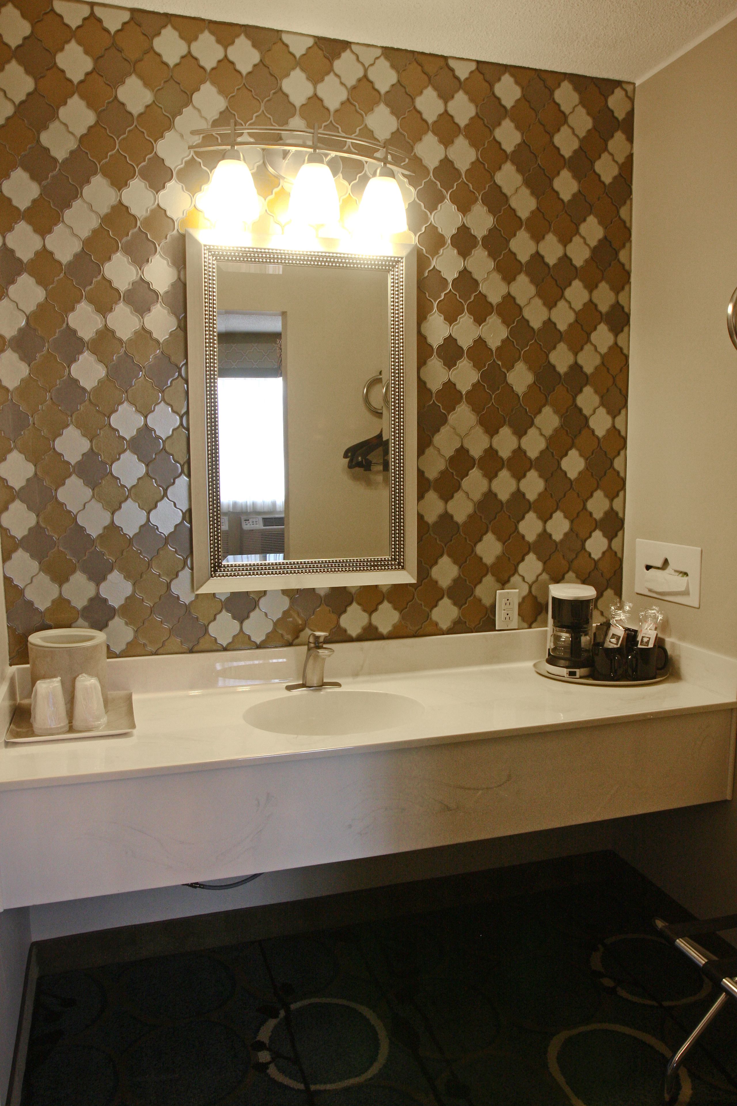 A bathroom with a white sink, a mirror with lights, a patterned wall, and a coffee maker on the counter.
