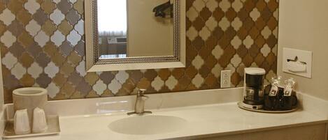 A bathroom with a white sink, a mirror with lights, a patterned wall, and a coffee maker on the counter.