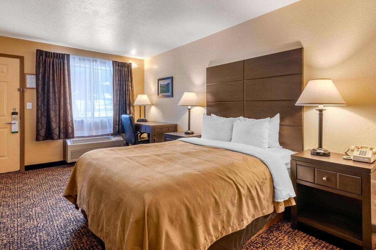 Een hotelkamer met een groot bed, twee bedlampen, een bureau met stoel, een raam met gordijnen en een thermostaat.