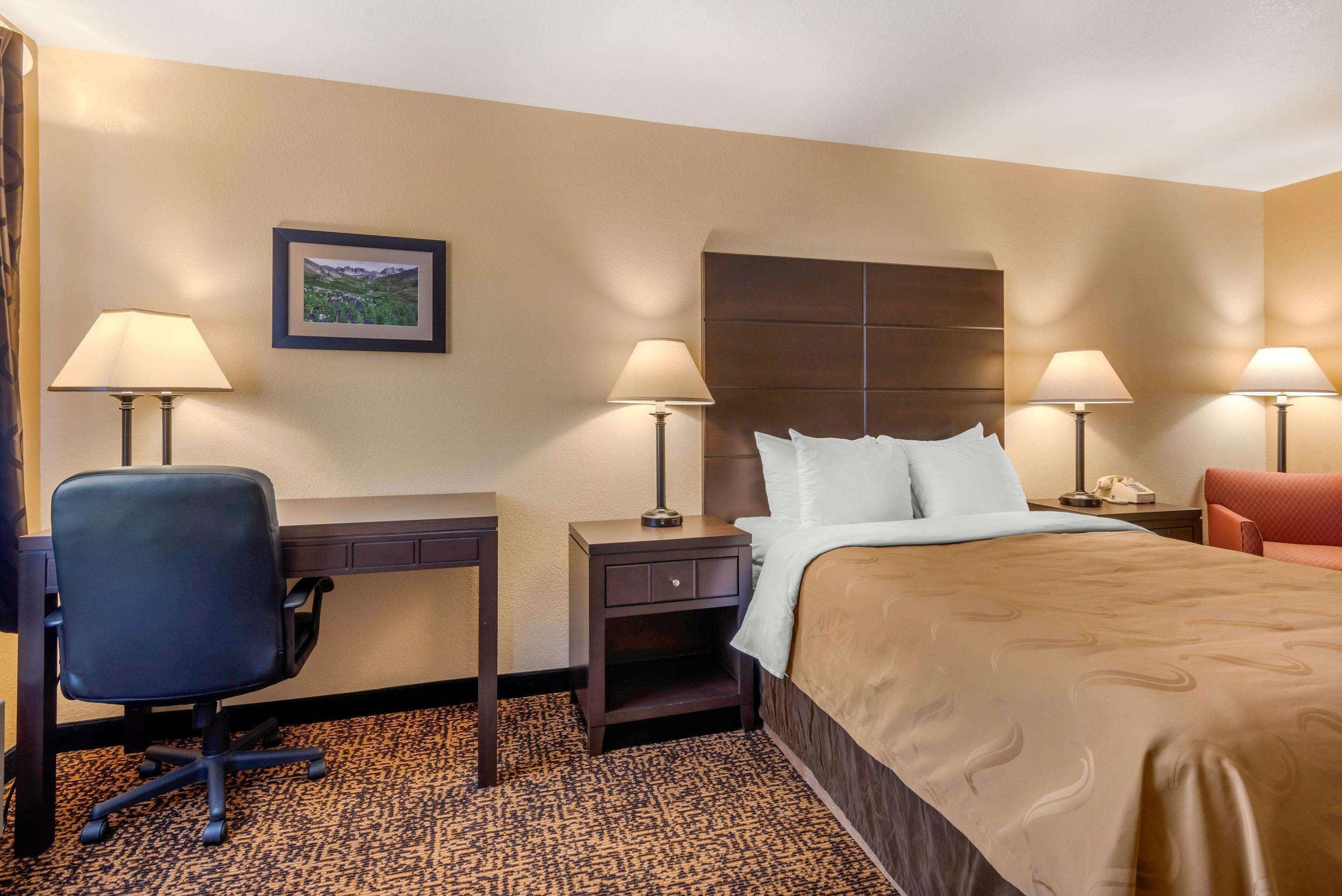 A hotel room with a bed, two bedside tables with lamps, a desk with a chair, and a framed picture on the wall.