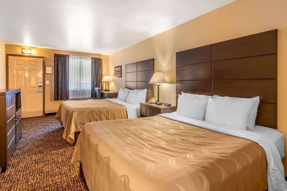 A hotel room with two beds, a desk, a chair, a lamp, and a door.