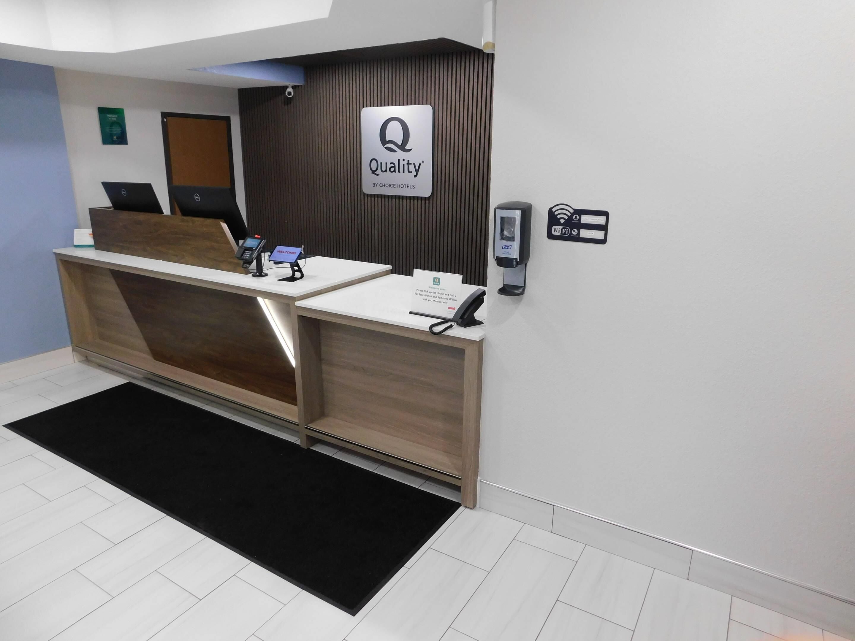 A hotel reception desk with a 'Quality by Choice Hotels' sign, a digital check-in kiosk, a phone, and a Wi-Fi sign.