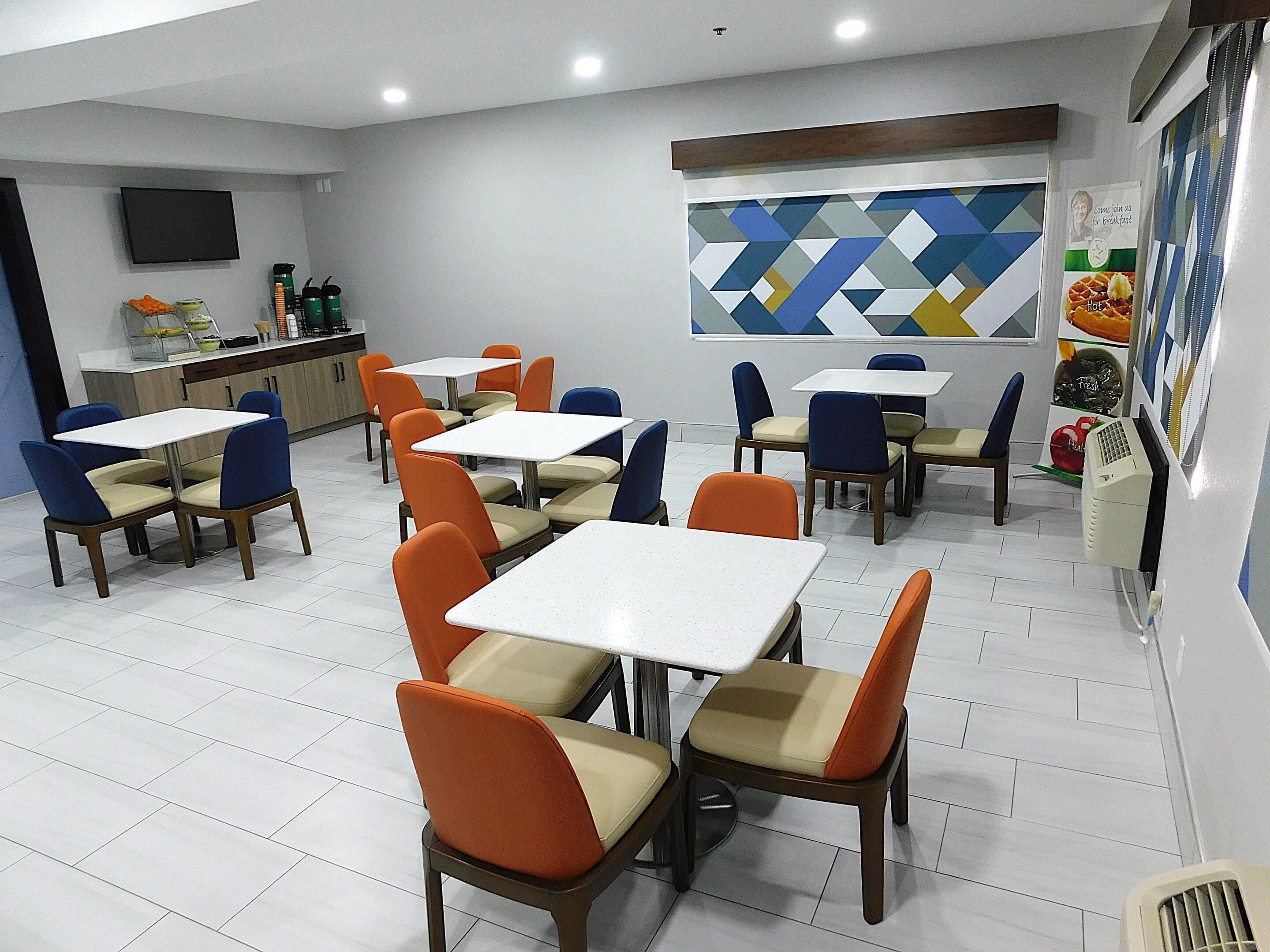 A restaurant interior with multiple tables and chairs, a kitchen area, and a wall with a geometric pattern.
